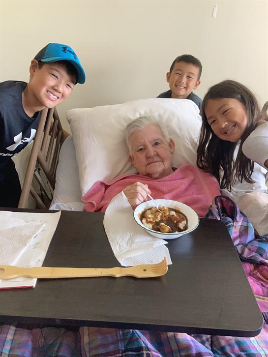 Four kids gather around a smiling elderly woman enjoying a bowl of dessert on a cozy afternoon.