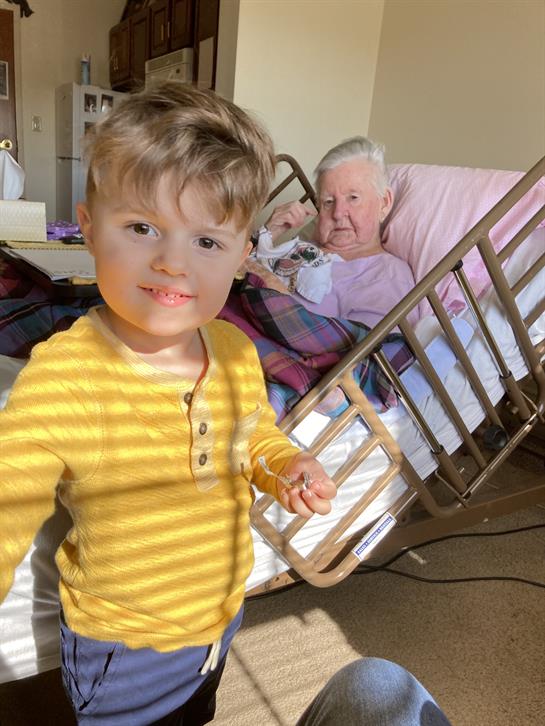 A young boy smiles at an elderly person seated in a bed, with sunlight streaming through the window.