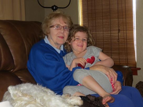 A grandmother and granddaughter smile together on the couch as a dog relaxes nearby.