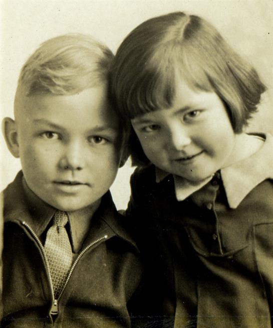 Two children pose side by side, sharing a warm smile, captured in a vintage setting.