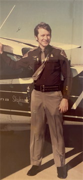 Law enforcement officer stands proudly next to a vintage police car in a rural setting.