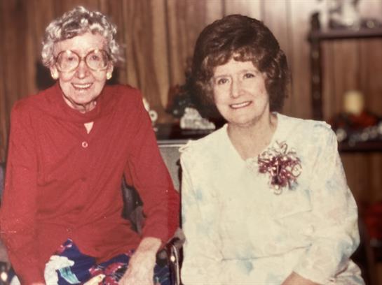 Two elderly women sit together, smiling warmly, in a comfortable living room setting.