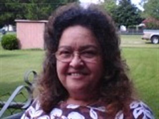 A woman with curly hair smiles while sitting outdoors in a garden filled with greenery and sunshine.