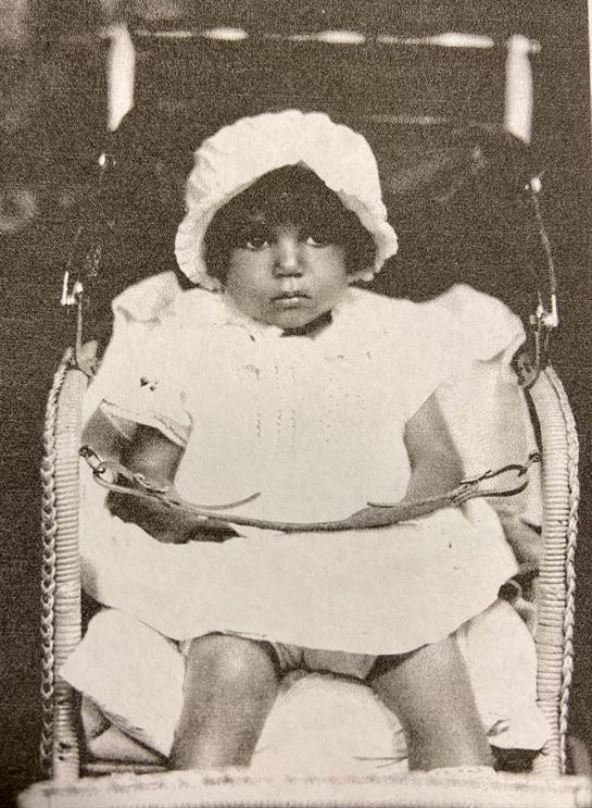 A child in a white dress and bonnet sits quietly in a charming chair, evoking the past.