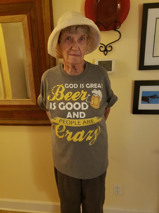 An elderly woman stands smiling in a vibrant gray shirt with playful text about beer and life.