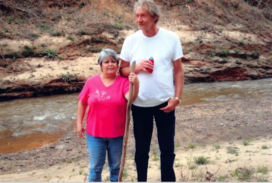 Two friends stand together by a river, surrounded by desert terrain, enjoying a sunny day outdoors.