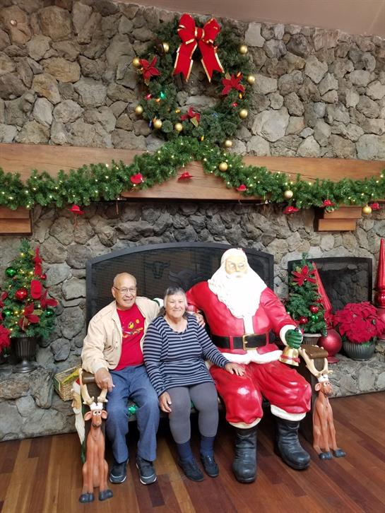 Two people pose with Santa Claus in a warmly decorated room with a stone fireplace.