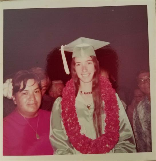 A proud graduate in a green gown and lei stands beside a family member during a ceremony.