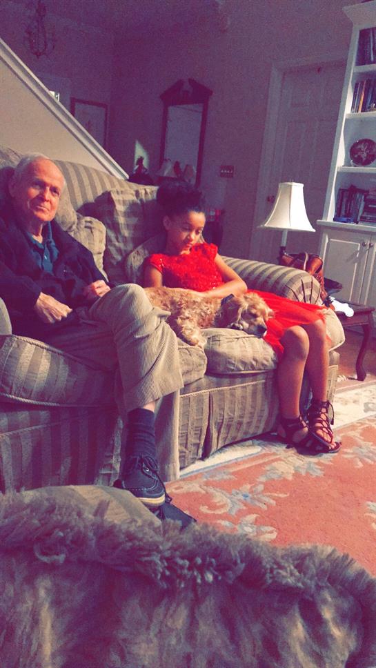 A man and a woman relax on a couch with a dog cuddled in between them in a warm living space.