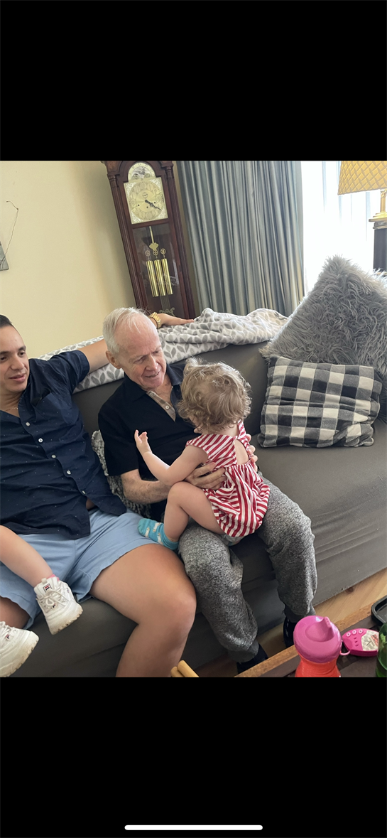Grandfather holding toddler while father relaxes on the couch in a warm, inviting living room.