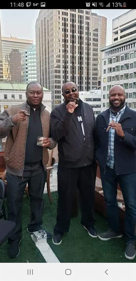 Three friends laugh and socialize while enjoying cigars and drinks on a rooftop.