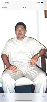 A man dressed in light clothing is seated indoors, looking calm and relaxed in an informal setting.