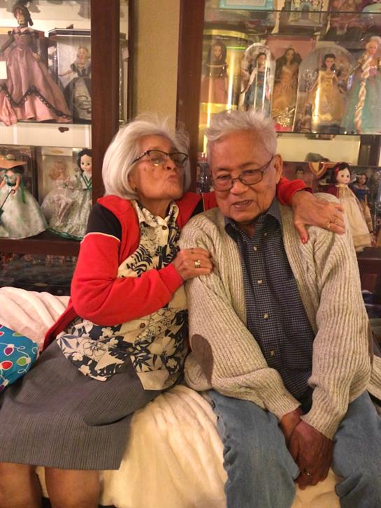 A loving elderly couple enjoys a tender moment in their home surrounded by cherished dolls.
