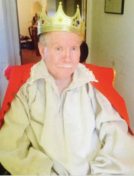 A senior man dressed in royal clothing with a golden crown sits proudly in a comfortable chair.