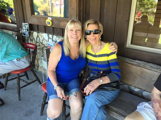 Two women smile while sitting on a bench outside a charming structure on a warm day.