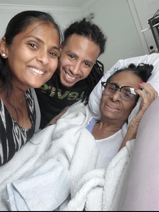 Two family members smile while visiting an elderly woman in a hospital bed.