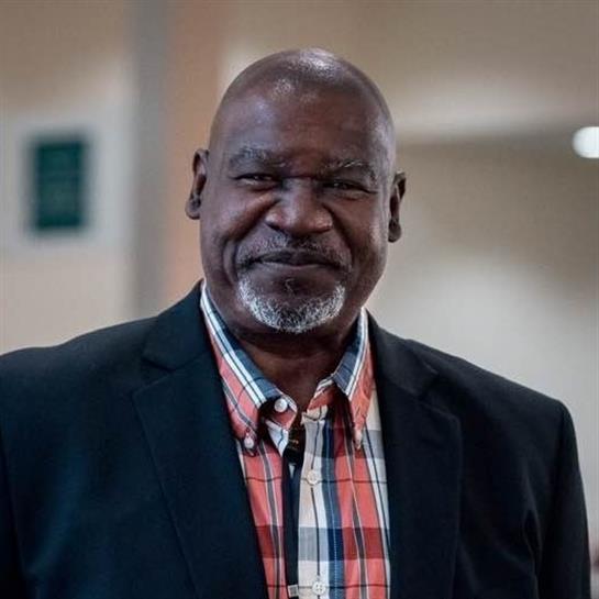 An older man stands confidently, wearing a black jacket and plaid shirt, smiling warmly indoors.