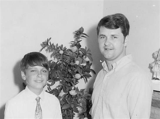 A young boy in a tie and an adult man pose happily near a decorative plant indoors.