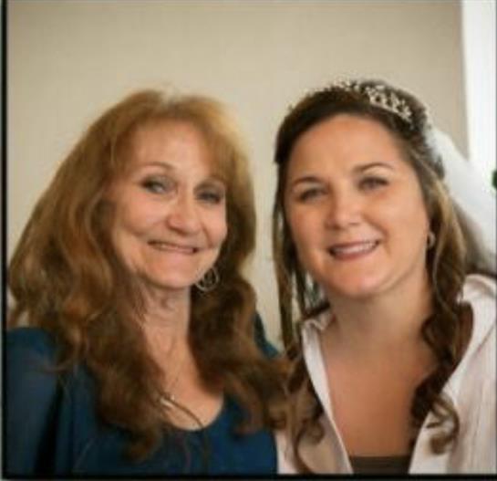 Two women share a joyful moment, smiling warmly while dressed for a festive occasion indoors.