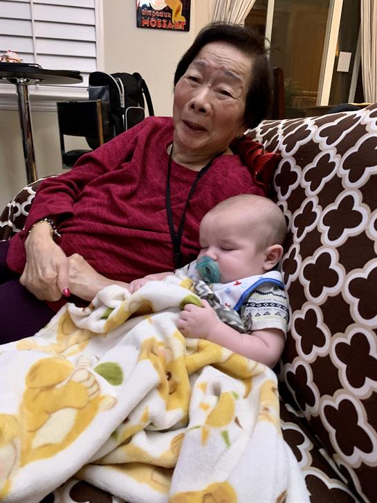 Grandmother smiles warmly while holding her sleeping grandchild on a soft couch.