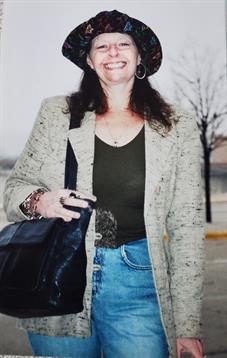 A woman smiles while wearing a stylish gray blazer and a hat, holding a bag outdoors.