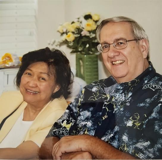 A happy couple sits close, smiling warmly in a bright, flower-filled room.