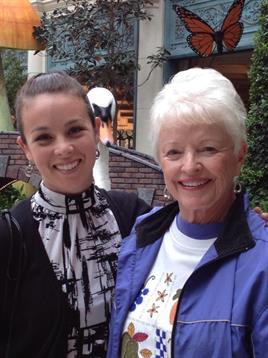 Two women stand side by side, smiling warmly in a garden setting with greenery and sunlight.