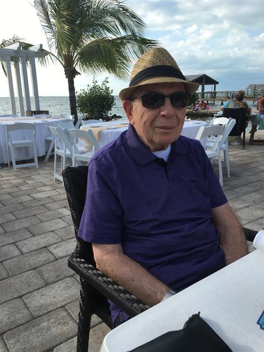 An elderly man enjoys the beachside view from an outdoor table, dressed casually with sunglasses.