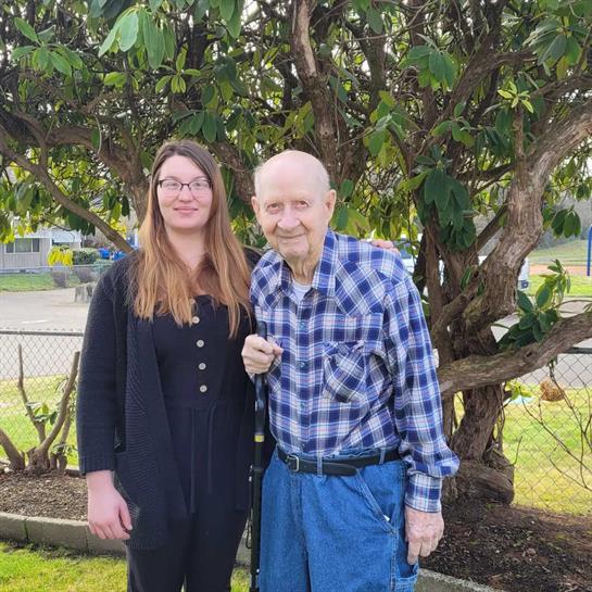 A young woman and an elderly man pose together by a vibrant tree on a warm, sunny day outside.