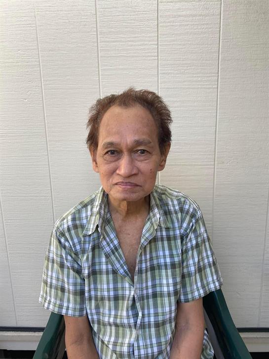 An elderly man with a neutral expression sits calmly on a chair, wearing a checkered shirt.