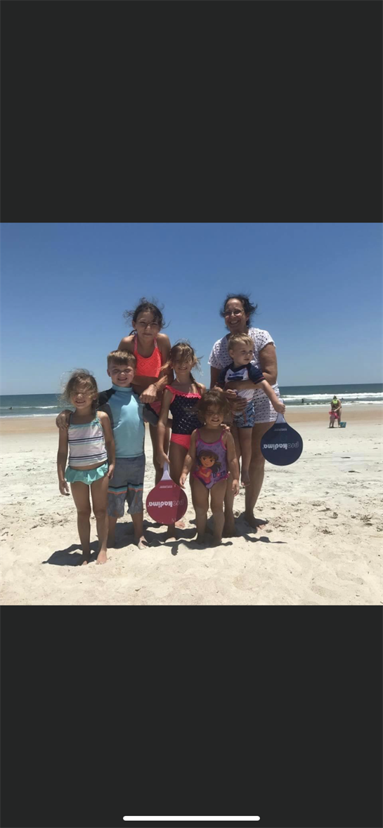 Seven children and one adult smile for a joyful group photo at the beach under sunny skies.