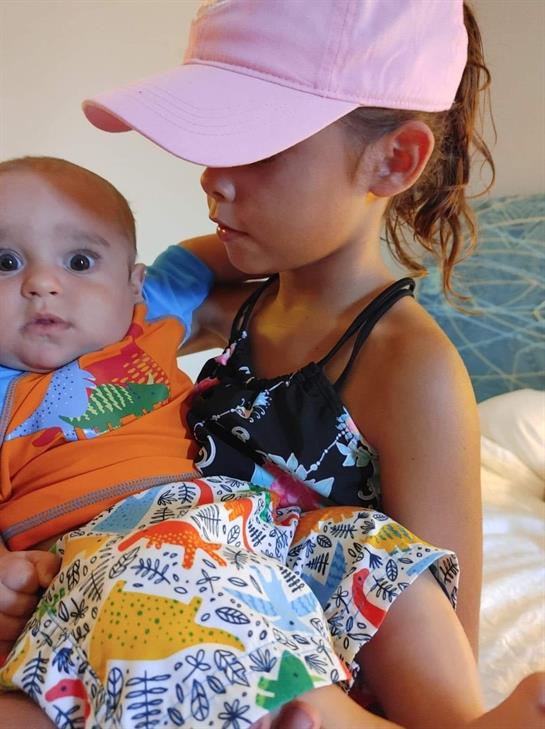 Young girl lovingly cradles her baby brother while both wear colorful attire indoors.