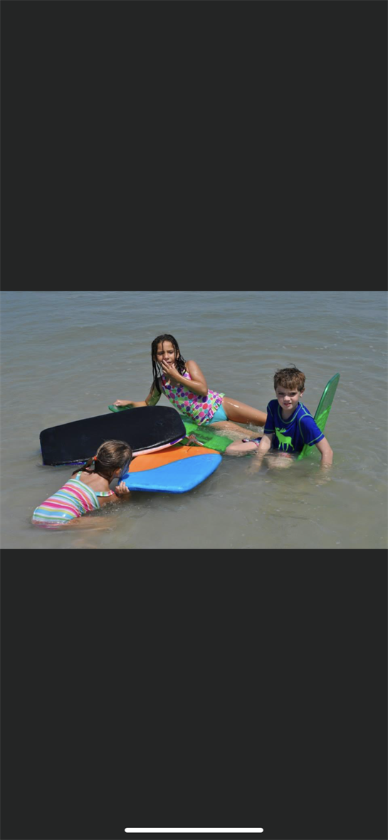 Three kids play with colorful boogie boards in shallow water on a sunny beach day.