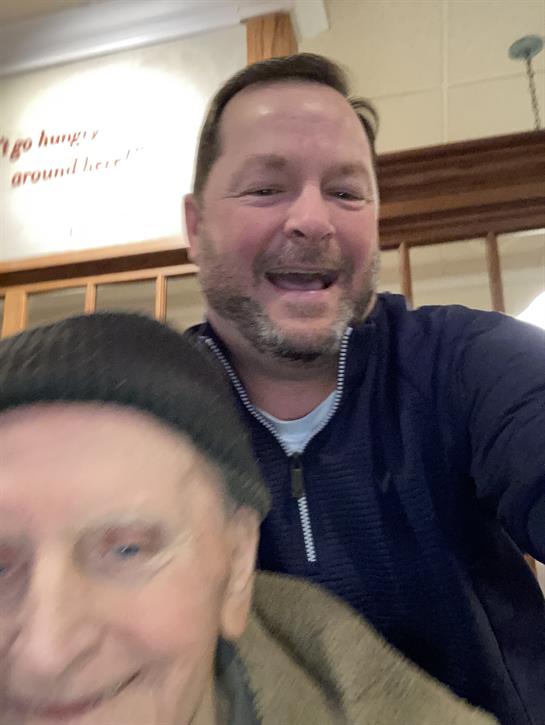 Two smiling men pose happily together in a warm and inviting cafe setting.