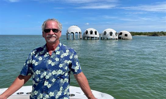 Man on boat in a bright shirt smiles at the camera as dome-shaped buildings rise from water.