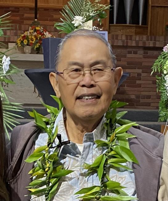 An elderly man wears a smiling expression with a traditional lei at an outdoor event.