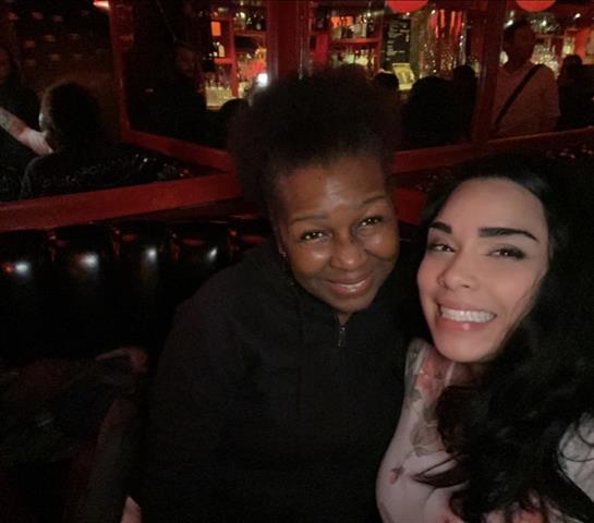 Two friends pose happily together in a vibrant bar setting filled with warm lights and reflections.