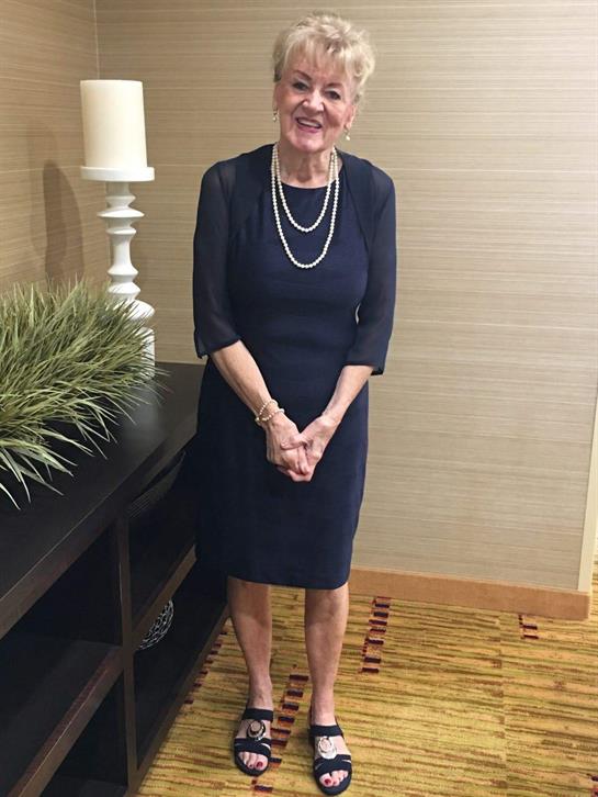 Smiling elderly woman wearing a stylish dress poses with her hands clasped beside greenery.