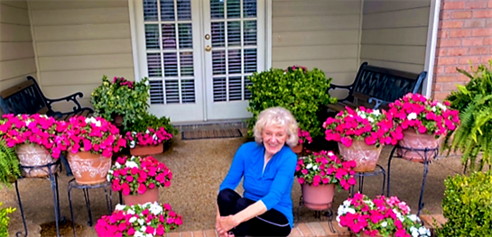 A woman kneels in her garden, smiling amid vibrant flowers, enjoying a sunny afternoon outdoors.