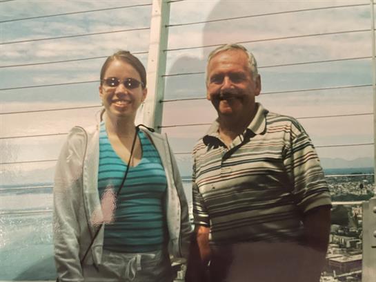 Two people pose happily outdoors, enjoying a sunny day by a rustic wooden building.