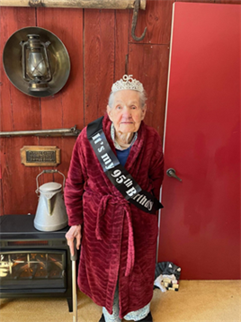 A woman dressed in a robe and tiara joyfully celebrates her 95th birthday while holding a cane.