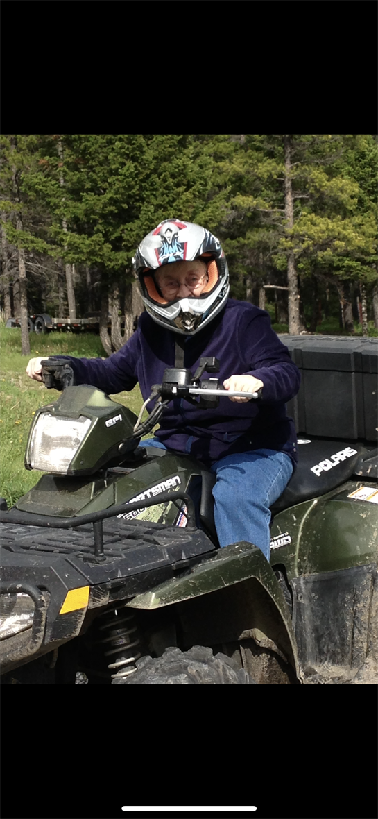 Older adult wearing protective gear operates an ATV on a grassy trail surrounded by trees.