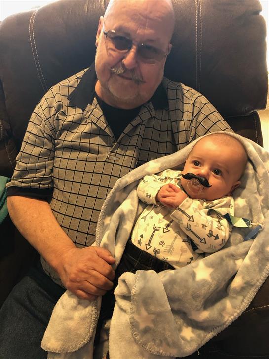 An elderly man and a baby wrapped in a blanket share playful smiles with mustache props.