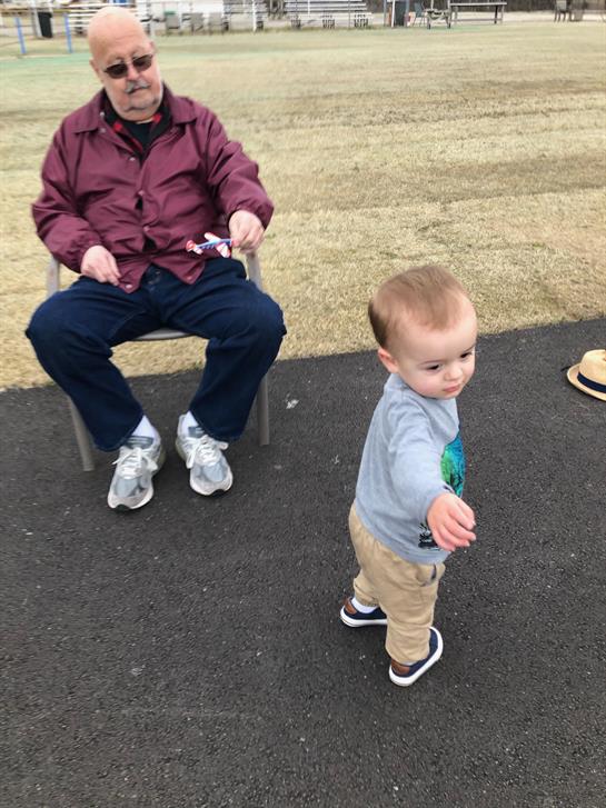 Elderly man enjoys a day in the park as a toddler walks around and explores the area.
