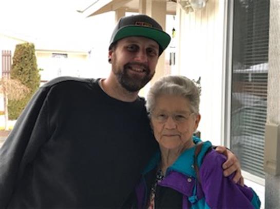 Elderly woman and adult man share a warm embrace outside a house under bright sunlight.