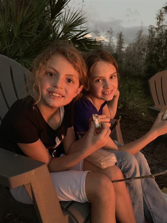 Two girls smile while roasting marshmallows around a campfire as the sun sets behind them.