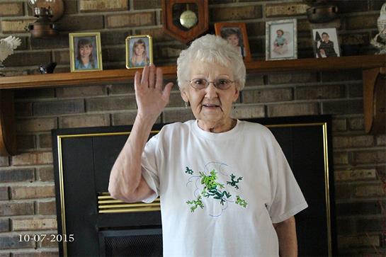 An elderly woman waves in a cozy living room filled with family portraits.