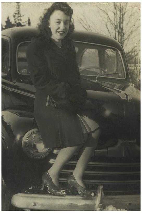 A woman dressed in a stylish outfit sits on a vintage car, exuding nostalgia in a cityscape.