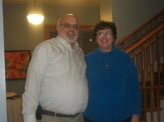 Two people smile warmly while standing side by side in a bright indoor space.