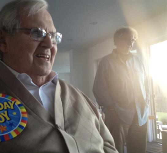 An elderly man enjoys a festive moment surrounded by family in a warm, light-filled room.